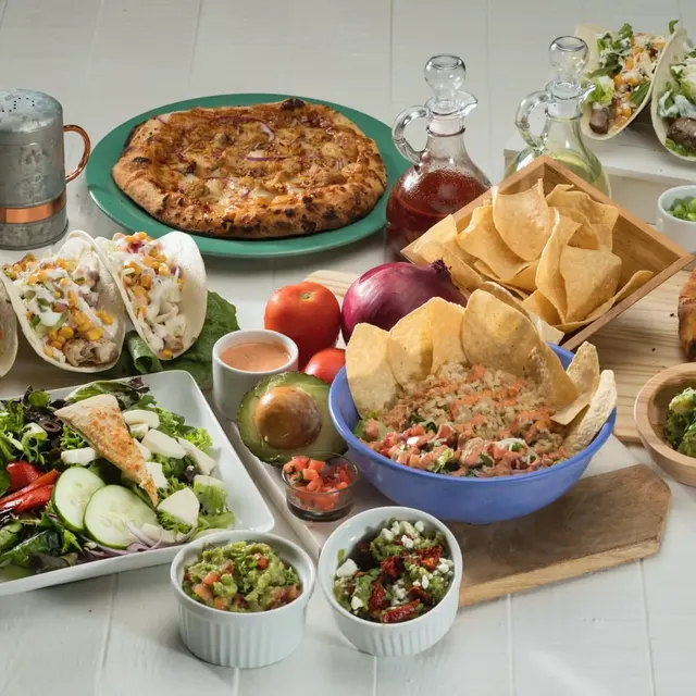 A spread of tacos, salad, chips, guacamole and two pizzas on a white table