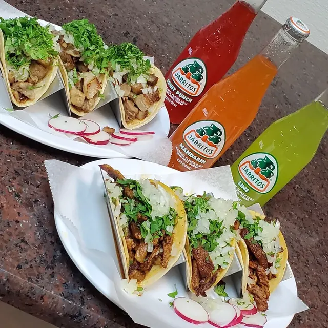 Two trays of tacos topped with cilantro and onion beside three bottles of Jarritos soda